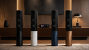 Four tall speakers in different colors arranged in a room with a television and lamp in the background.